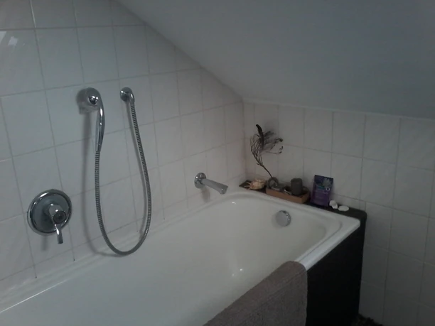 Ferienwohnung Carpe Diem Bad Badewanne mit Duschkopf in einem Badezimmer mit weiß gefliester Schräge und minimalistischem Dekor.