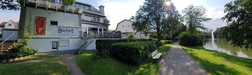 Außenansicht Das Restaurant "Blaues", ein Gebäude mit Terrasse, liegt direkt an einem ruhigen Flussweg umgeben von Bäumen.