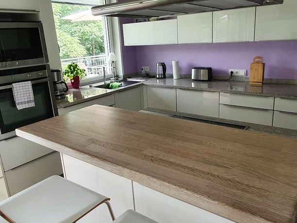 Moderne Küche mit großer Holzarbeitsfläche, weißen Schränken und lila Wand, Blick ins Grüne.