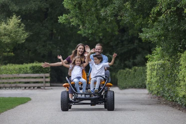 Gocart Giebner in Monheim am Rhein Eine Familie fährt auf einem viersitzigen, orangenen Gocart fröhlich auf einer von Bäumen gesäumten Straße.