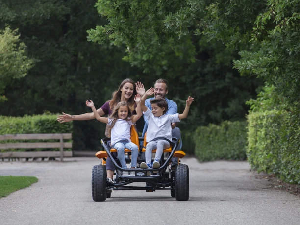 Gocart Giebner in Monheim am Rhein Eine Familie fährt auf einem viersitzigen, orangenen Gocart fröhlich auf einer von Bäumen gesäumten Straße.