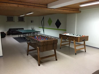 Ein Raum mit Tischfußballtisch und Tischtennisplatte, umgeben von weißen Wänden und Holzdecke.