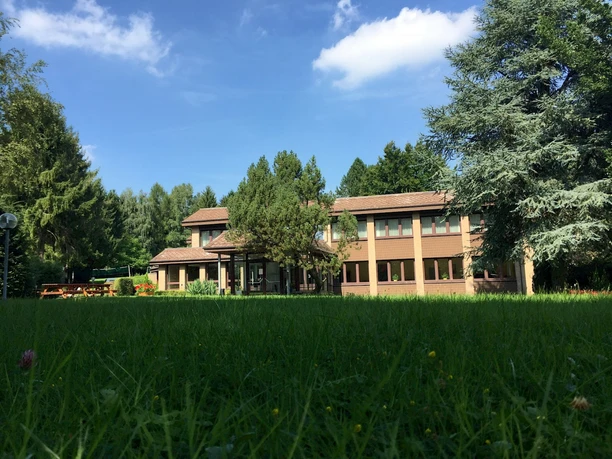 Backsteinhaus mit Flachdach, umgeben von Bäumen und Rasen unter blauem Himmel mit vereinzelten Wolken.