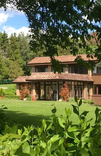 Großes zweistöckiges Haus mit großen Fenstern, umgeben von grünen Bäumen und einer gepflegten Wiese.