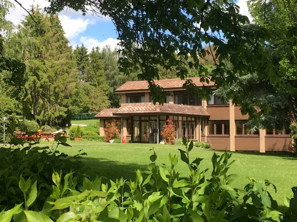 Großes zweistöckiges Haus mit großen Fenstern, umgeben von grünen Bäumen und einer gepflegten Wiese.