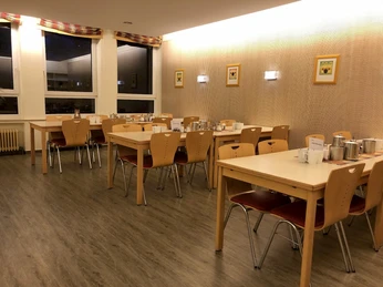 Ein heller Speisesaal mit Holzstühlen und Tischen, gedeckt für mehrere Gäste, mit Wanddekoration.