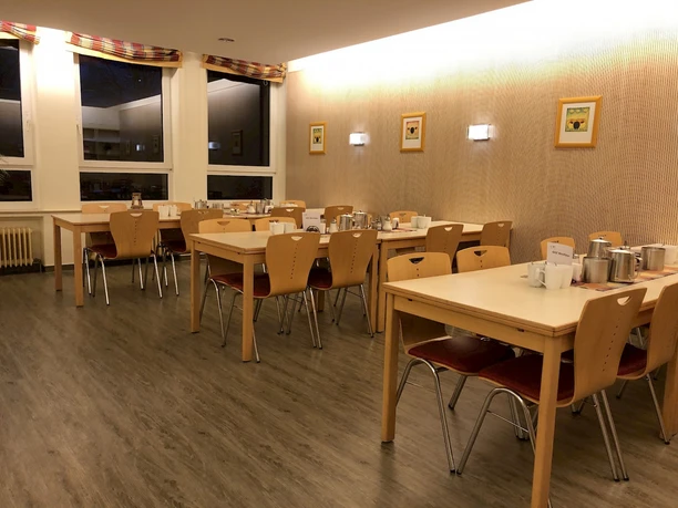 Speisesaal Ein heller Speisesaal mit Holzstühlen und Tischen, gedeckt für mehrere Gäste, mit Wanddekoration.