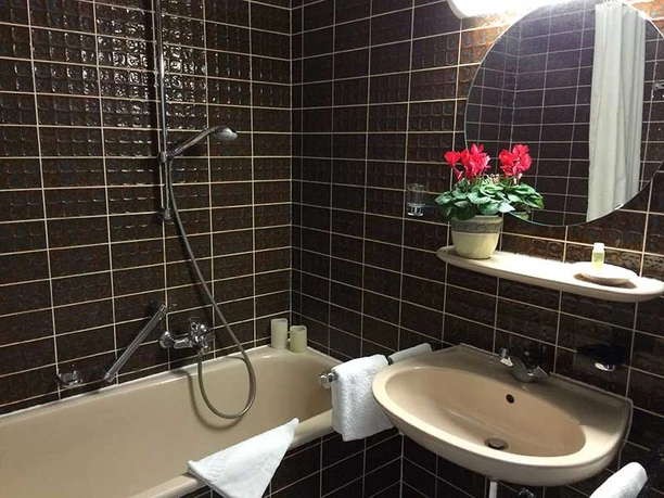 Bad.jpg Bad mit dunklen Fliesen, ovalem Spiegel, Waschbecken und Wanne; rote Blume auf Ablage.