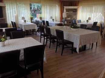 Speisesaal.jpg Ein lichtdurchfluteter Speisesaal mit gedeckten Tischen und schwarzen Stühlen, umgeben von großen Fenstern.