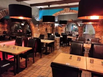 Essbereich in einem Restaurant. Man sieht gefliesten Boden und gemauerte Wände. In der Mitte des Raumes sind Tische und Stühle. Von der Decke hängen Lampen mit schwarzem Lampenschirm