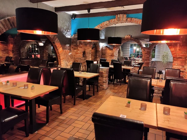 Essbereich in einem Restaurant. Man sieht gefliesten Boden und gemauerte Wände. In der Mitte des Raumes sind Tische und Stühle. Von der Decke hängen Lampen mit schwarzem Lampenschirm