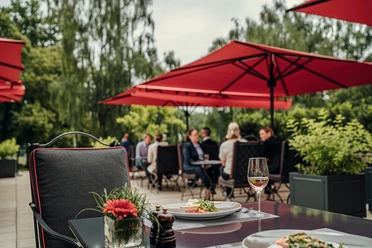 Auf der Außenterrasse des Bistrots Quellenhof befinden sich mehrere Sitzgruppen mit Personen. Sie werden von roten, quadratischen Schirmen überdacht. Im Vordergrund steht ein mit Essen und Gläsern gedeckter Tisch und ein leerer Stoffstuhl.Im Hintergrund stehen Bäume.