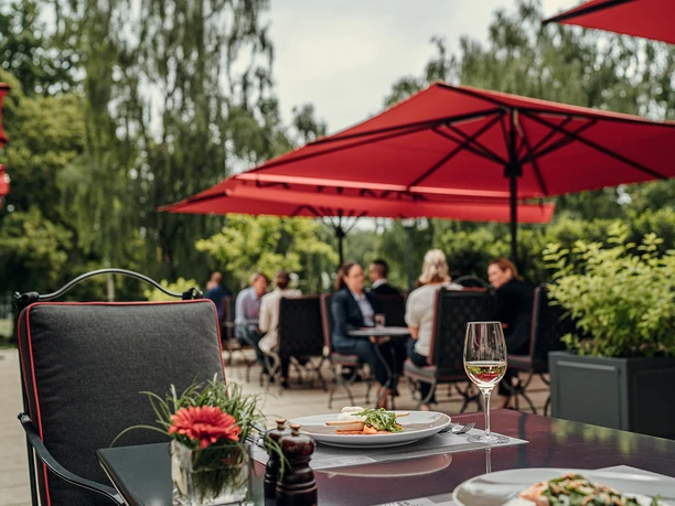 Auf der Außenterrasse des Bistrots Quellenhof befinden sich mehrere Sitzgruppen mit Personen. Sie werden von roten, quadratischen Schirmen überdacht. Im Vordergrund steht ein mit Essen und Gläsern gedeckter Tisch und ein leerer Stoffstuhl.Im Hintergrund stehen Bäume.