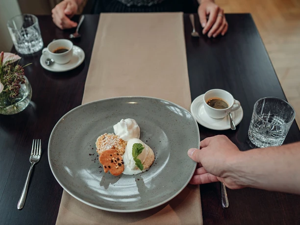 Bistro Quellenhof Auf einem Tisch liegt ein heller, länglicher Tischläufer und Gedeck für zwei Personen, das Wassergläser, Espressos und Besteck umfasst. Eine Hand stellt gerade einen Teller mit Dessert auf den Tischläufer.