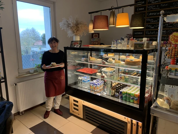 Auslage in einem Café. Man sieht eine Frau, die einen Teller mit einem belegten Brötchen hält. Neben ihr ist eine Vitrine mit Kuchen, Limonade und Säften.