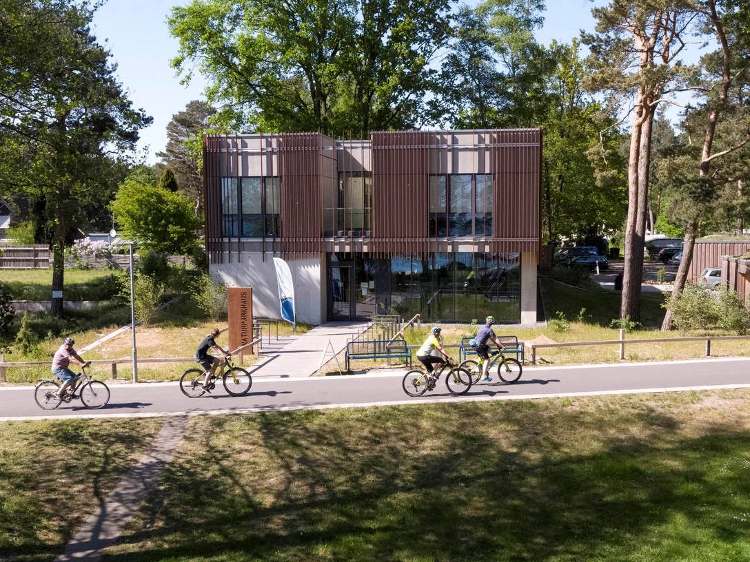 Naturpark-Haus Das Naturpark-Haus mit seiner modernen Glasfassade ist von Radfahrern auf einer Allee umgeben.The Nature Park House with its modern glass façade is surrounded by cyclists on an avenue.Naturparkhuset med sin moderne glasfacade er omgivet af cyklister på en allé.Het Natuurparkhuis met zijn moderne glazen gevel wordt omringd door fietsers op een laan.