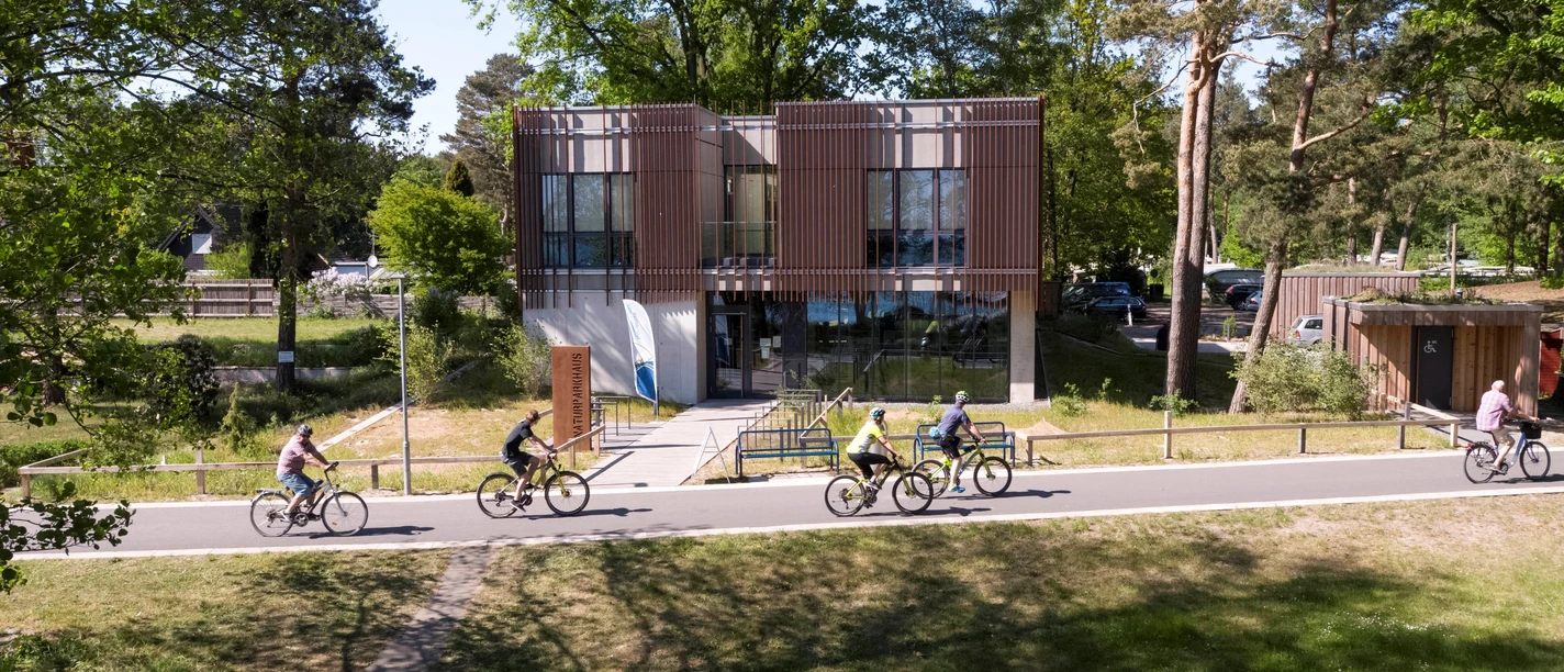 Naturpark-Haus The Nature Park House with its modern glass façade is surrounded by cyclists on an avenue.
