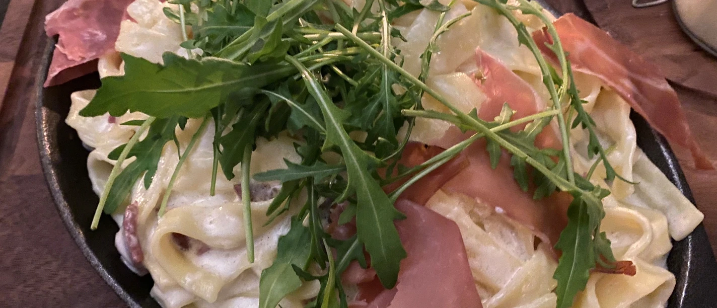 Nudelgericht Ein Teller mit Tagliatelle in cremiger Soße, garniert mit Rucola und Schinken auf einem Holztisch.
