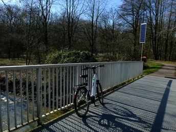 E-Bike am Meschesee.JPG Ein E-Bike steht auf einer Brücke über einen Bach im Wald, daneben ein Solarpanel.