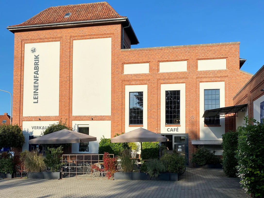 Backsteingebäude mit grauer Fassade, schattiger Terrasse, Verkaufsraum und Café unter Sonnenschirmen.