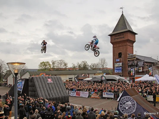Stuntshow in Augustfehn Motorradstuntfahrer springen durch die Luft vor einer großen Menschenmenge am Eisenhüttenplatz.