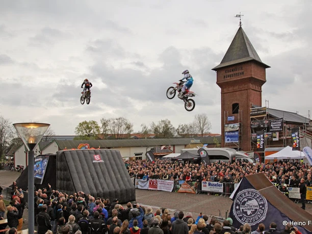 Stuntshow in Augustfehn Motorradstuntfahrer springen durch die Luft vor einer großen Menschenmenge am Eisenhüttenplatz.