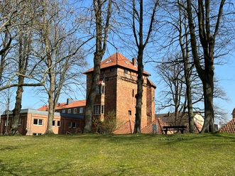 Backstein-Turm mit roten Ziegeldächern, umgeben von kahlen Bäumen, vor blauem Himmel im Hintergrund.
