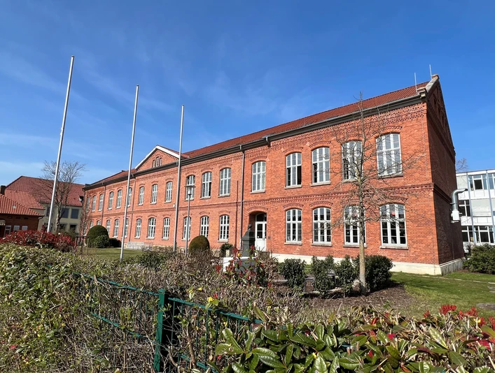 Polizeiakademie Backsteinbau der Polizeiakademie mit großen Fenstern, umgeben von gepflegten Pflanzen und leeren Fahnenmasten.