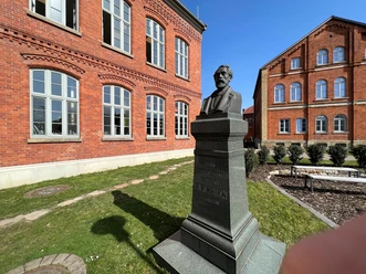 Rhien-Büste Büste des Heinrich Hermann Reineke vor einer roten Backsteinfassade in grünem Außenbereich.