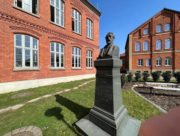 Rhien-Büste Büste des Heinrich Hermann Reineke vor einer roten Backsteinfassade in grünem Außenbereich.