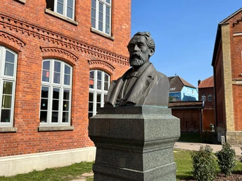 Rhien-Büste Bronzebüste eines Mannes auf einem steinernen Sockel vor einem Backsteingebäude mit weißen Fenstern.