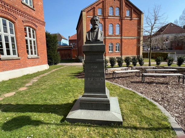 Rhien-Büste Eine Büste von Simon Rhenius auf einem Sockel in einem grünen Innenhof mit Backsteingebäuden.