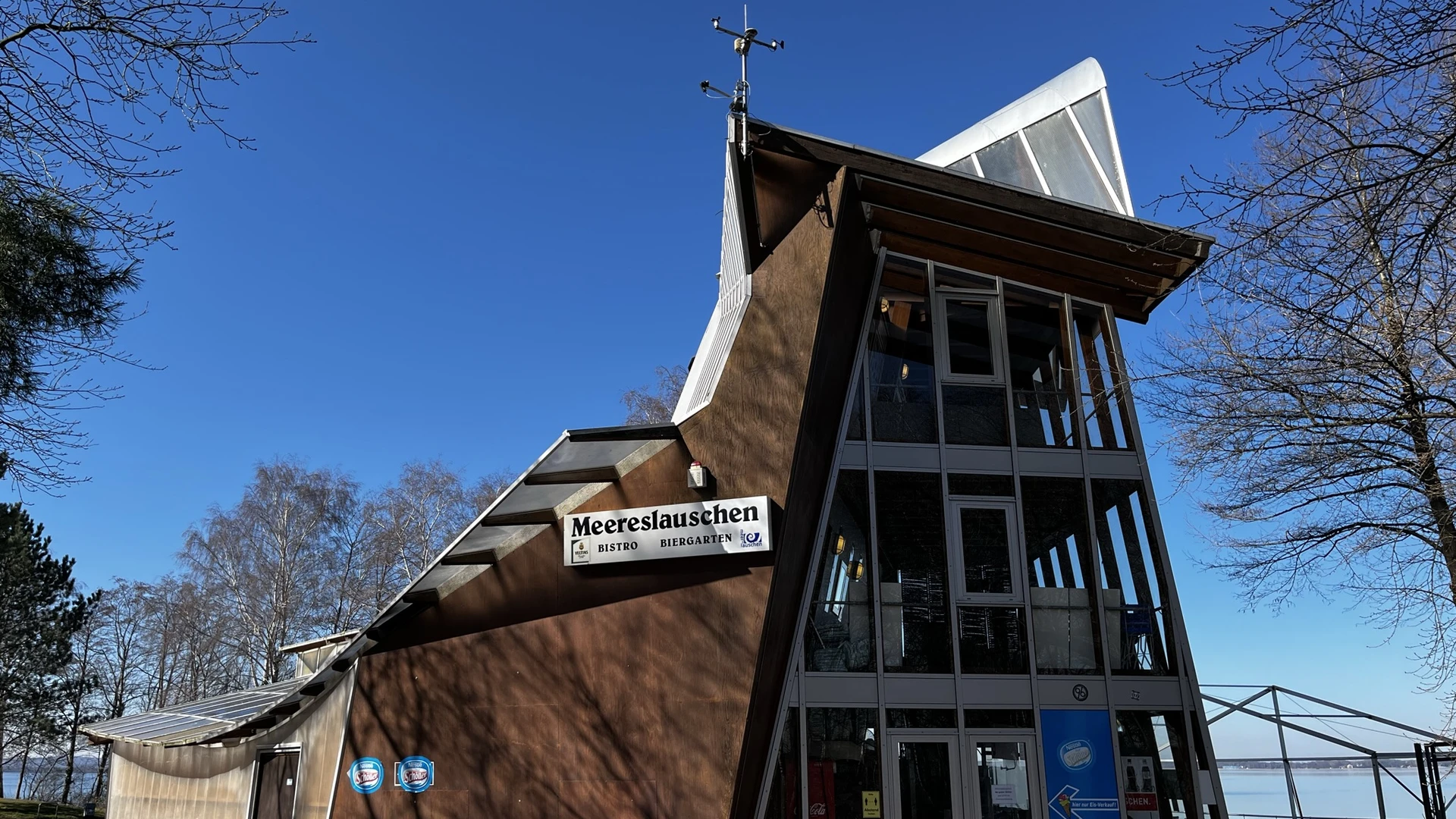 Badeinsel Steinhude - Bistro Meereslauschen Modernes Bistro mit markanter Architektur, Glasfassade und großem Namensschild, umgeben von Bäumen.