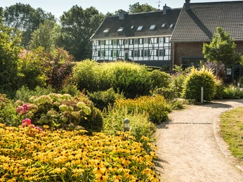 Hofcafé Abtsküche à Heiligenhaus Un chemin mène à travers un jardin luxuriant jusqu'à une maison à colombages sous un ciel clair.