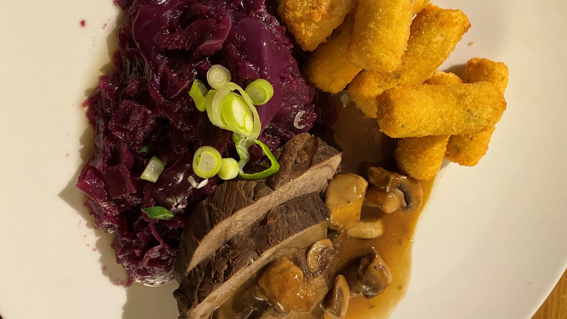 Roast venison with glazed mushrooms, served with red cabbage and crispy croquettes on a white plate.