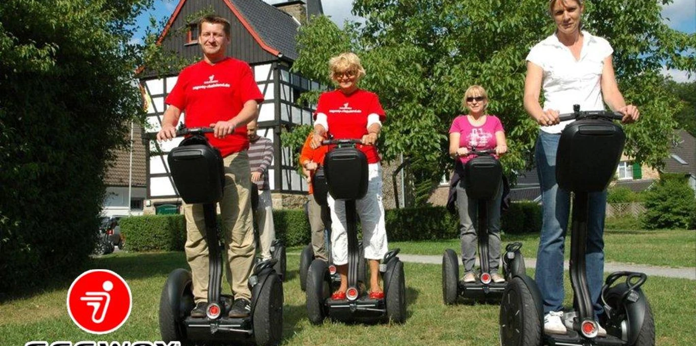 SEGWAYPoint-Haan-Renania Quattro persone percorrono allegramente a bordo di un Segway un sentiero verde di fronte a una casa a graticcio.