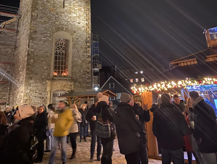 Noel Pazarı2021_Platz Am Offers_StadtVelbert_Schuster_image00036.jpeg Akşam ışığında, kuleli ışıklandırılmış bir kilisenin etrafındaki Noel pazarının tadını çıkarıyorlar.