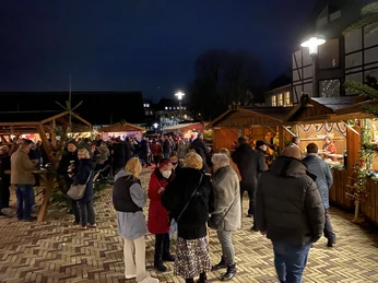 Noel Pazarı2021_Platz Am Offers_StadtVelbert_Schuster_image00048.jpeg İnsanlar, şenlikli bir şekilde dekore edilmiş ahşap tezgahların bulunduğu akşam Noel pazarında geziniyor.