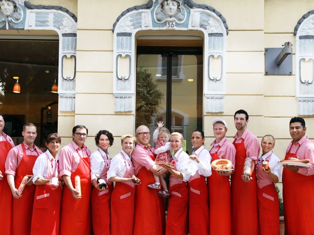Macellaio Rauschmann a Haan-Gruiten Un gruppo di persone con grembiuli rossi sorride davanti a un edificio con le finestre decorate.