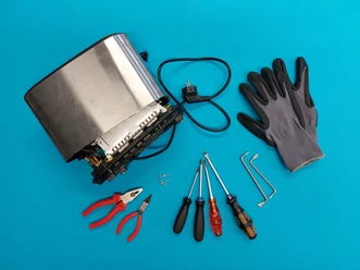 Repair Cafe, maar dan andersom. Gedemonteerde broodrooster met gereedschap en handschoenen, klaar voor reparatie, op een blauwe achtergrond.