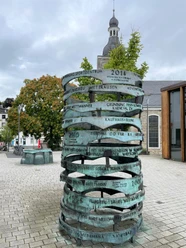 Herinneringen in Mettmann Een bronzen zuil met gegraveerde data staat voor een kerk onder een bewolkte hemel.