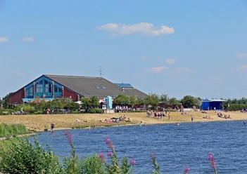 VVV-kantoor van de gemeente Geeste Mensen ontspannen aan de oever van het meer voor een groot rood gebouw met een café en bloeiende planten.