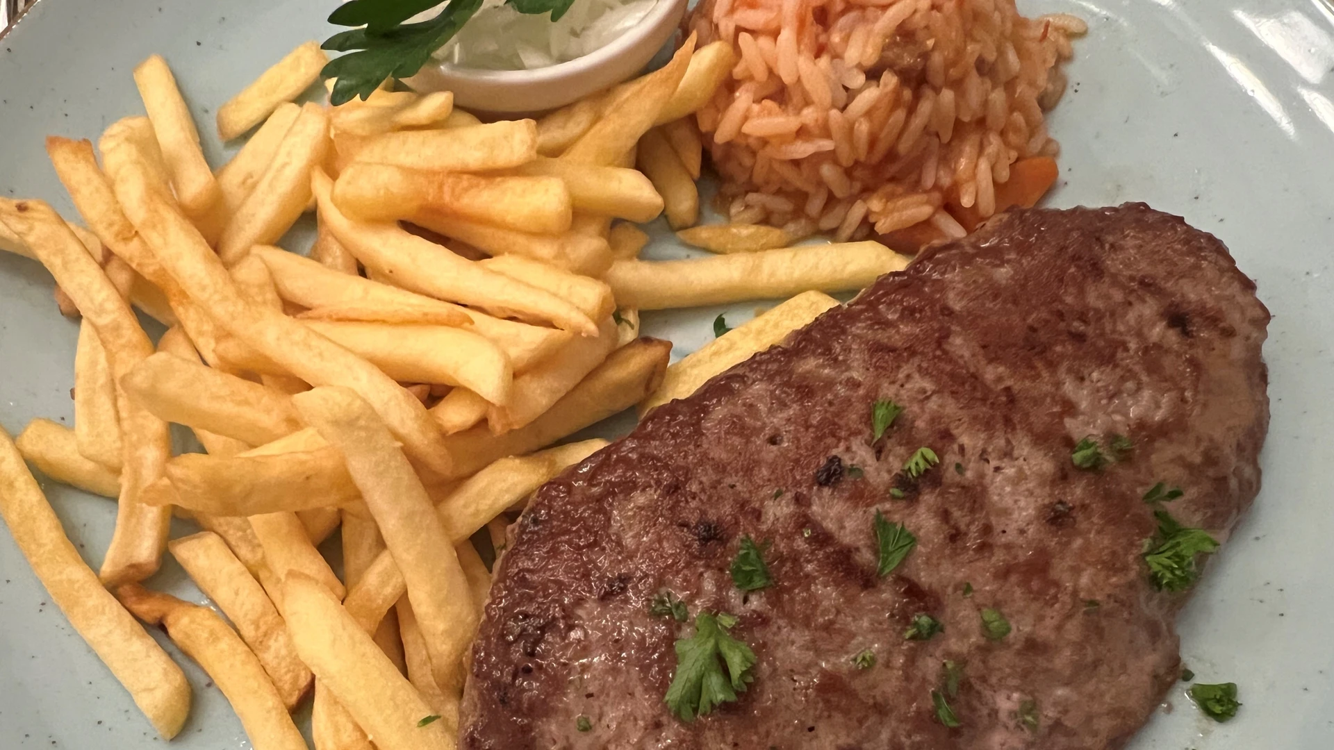 A juicy minced steak with parsley garnish, French fries and tomato rice on a blue ceramic plate.