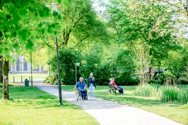 Wandeling in Altenberg Een rolstoelgebruiker wordt begeleid door een vrouw op een zonnig parkpad, terwijl een andere persoon op een bankje zit.