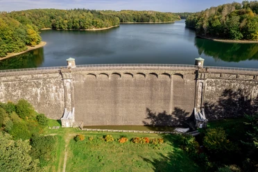 Neyetal Dam Dam in an idyllic landscape, surrounded by forest and water, under a blue sky.