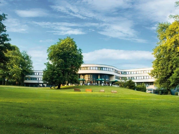 Best Western Plus Parkhotel Velbert Nowoczesny budynek hotelowy otoczony jest przestronnym, zielonym parkiem z ławkami.