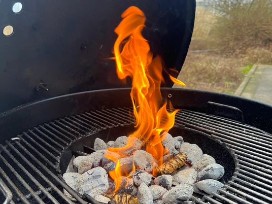 Fiamma nella griglia del bollitore Una fiamma alta, di colore giallo-arancio, si alza da una griglia piena di carboni ardenti.