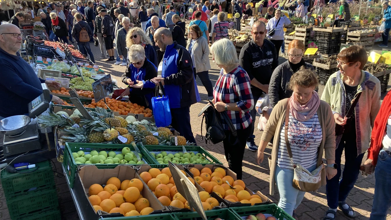 Markt mit vielen Menschen, die Obst und Gemüse kaufen. Im Vordergrund sind grüne Kisten mit Äpfeln, Orangen und Äpfeln. Im Hintergrund sind Stände mit weiteren Lebensmitteln.