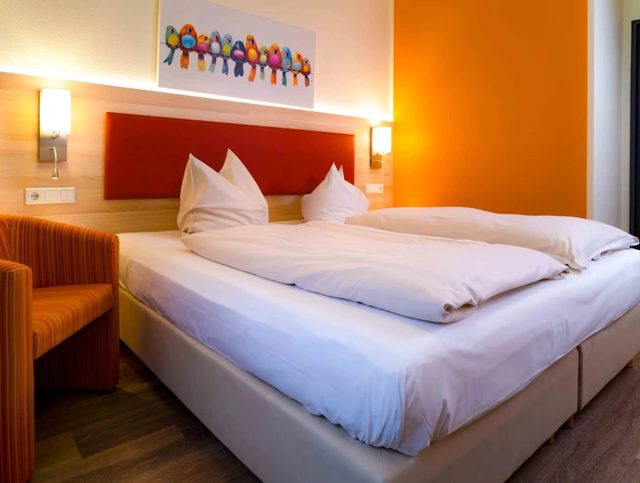 Pokój dwuosobowy w hotelu am Stadtpark Dwa jasne pojedyncze łóżka, pomarańczowe akcenty w całym pokoju, w tym kolorowy obraz przedstawiający ptaka na ścianie.