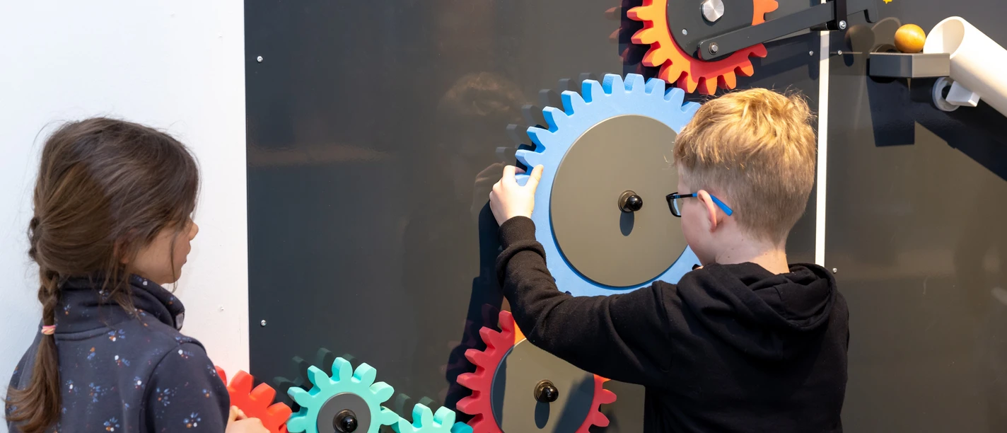 Chain reaction for families Two children interact with colorful gears on a blackboard, which represents an experimental field.